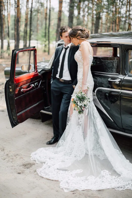 A newlywed couple poses next to a vintage black car in a woodland setting, with the bride holding a bouquet and showcasing her elaborate lace wedding dress and long train.