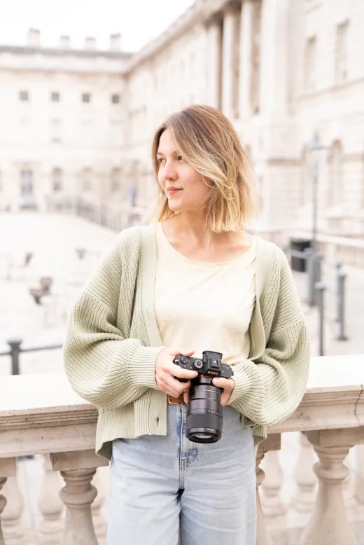 A woman with a bob haircut wearing a sage green cardigan holds a professional camera while standing outdoors against a classic architectural backdrop.