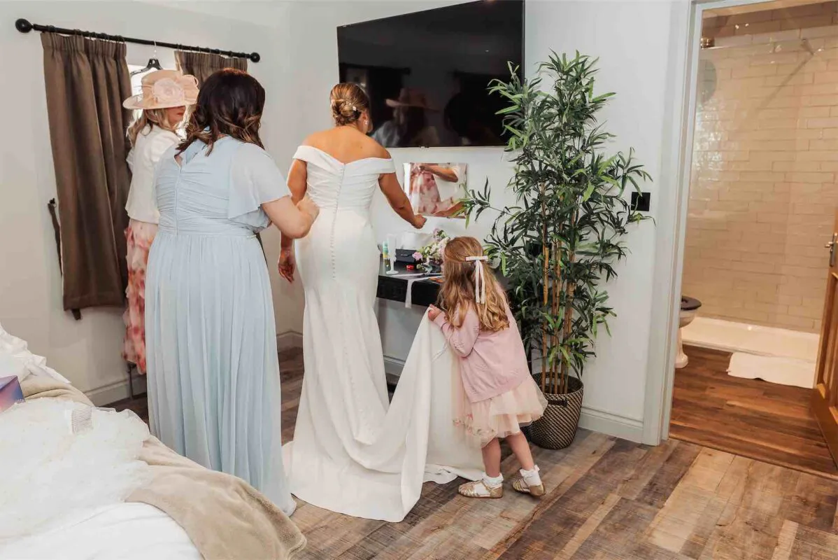 A bride in an off-the-shoulder gown with a button-back design stands with her bridesmaids and a flower girl helping with the train during morning preparations.