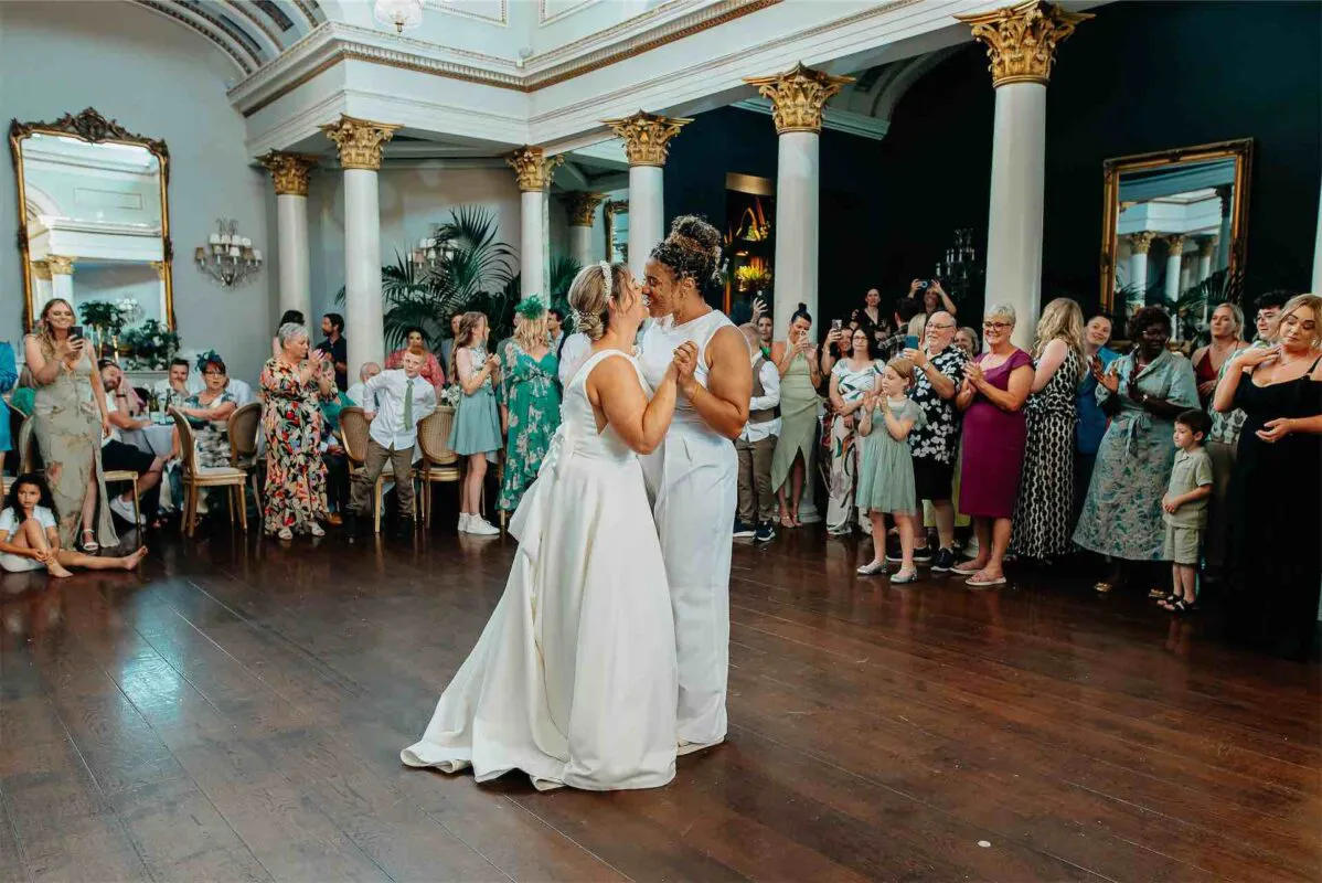 Two brides share a tender first dance together in a grand ballroom surrounded by smiling wedding guests.