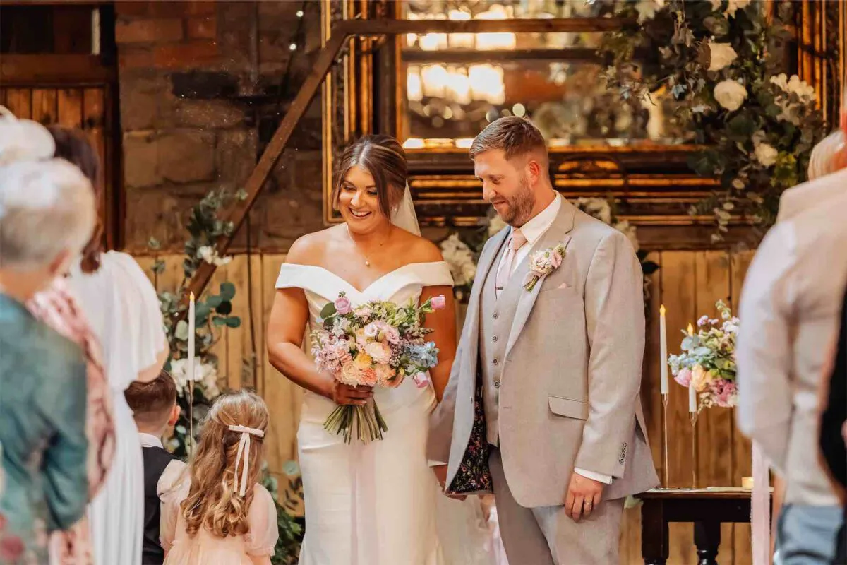 A smiling bride and groom stand together holding hands during their wedding ceremony inside a rustic barn venue with exposed brick walls.