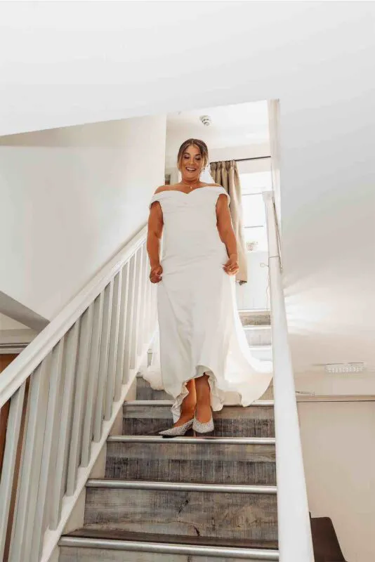 A smiling bride wearing an off-the-shoulder white crepe wedding dress walks down a wooden staircase during morning preparations.