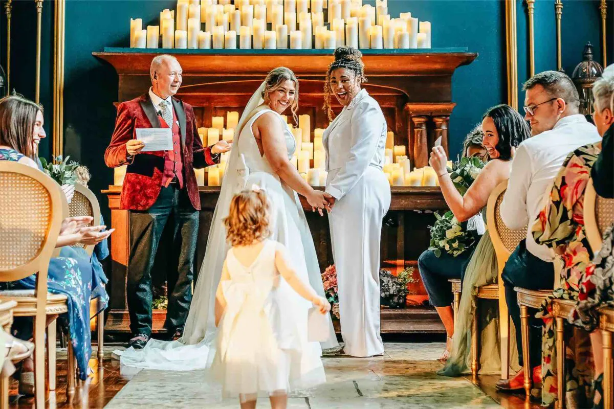 Two smiling brides hold hands at the altar during a candlelit ceremony, while a flower girl walks down the aisle towards them and guests cheer.
