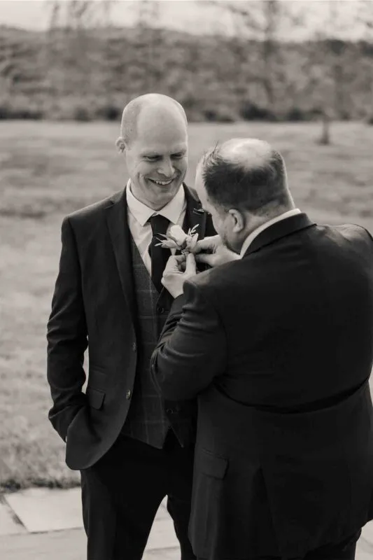 A groomsman proudly attaches a delicate boutonniere to the lapel of a smiling groom wearing a tweed waistcoat outdoors.