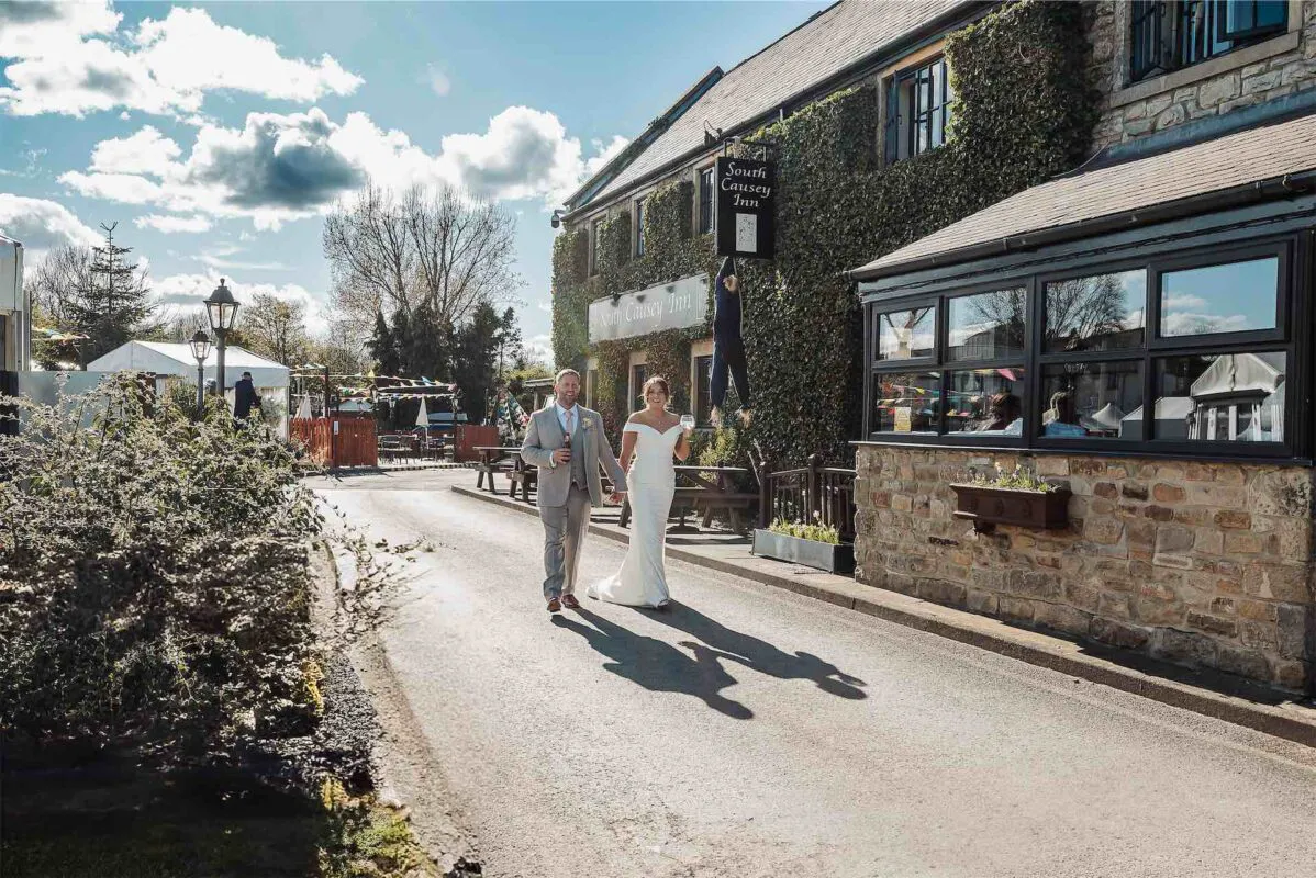 A newlywed couple walk hand in hand down a sunny pathway outside the South Causey Inn, laughing together on their wedding day.