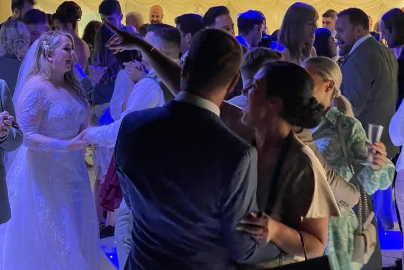 A joyous wedding reception in a marquee where guests laugh and dance under vibrant blue lighting on the packed dance floor.