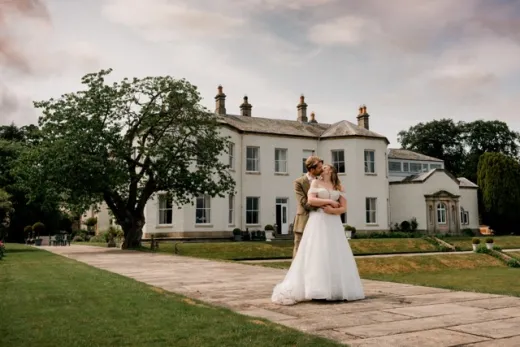 A newlywed couple shares a romantic kiss on the stone paved path in front of a grand country house manor.