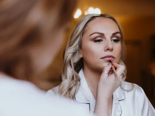 A close-up shot captures a blonde bride having her lipstick carefully applied by a makeup artist during morning bridal preparations.