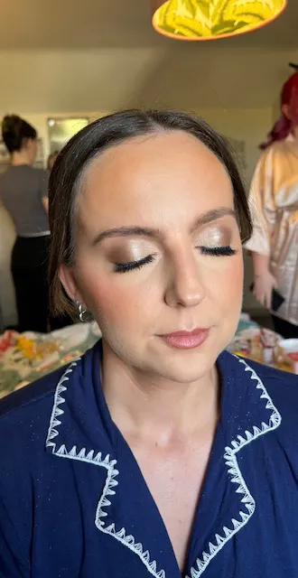 A close up portrait of a bride getting ready with her eyes closed, showcasing soft glam makeup and her hair styled in a low bun while wearing a dark blue patterned robe.