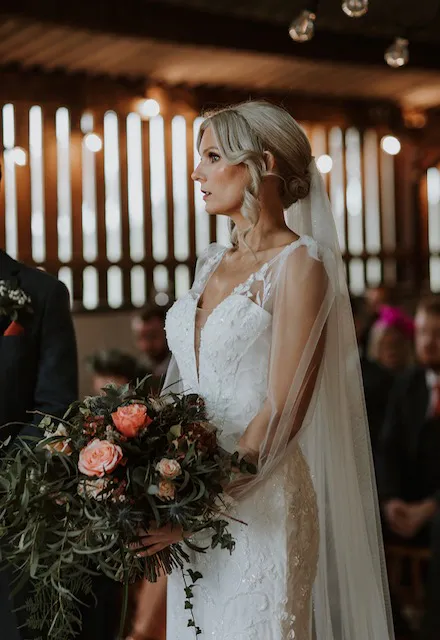 A profile shot of a bride holding a lush wild-style bouquet while standing inside a rustic wooden barn during her wedding ceremony.