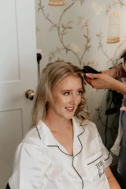A smiling bride wearing white satin pyjamas gets her blonde hair styled into soft waves by a hairstylist during wedding morning preparations.