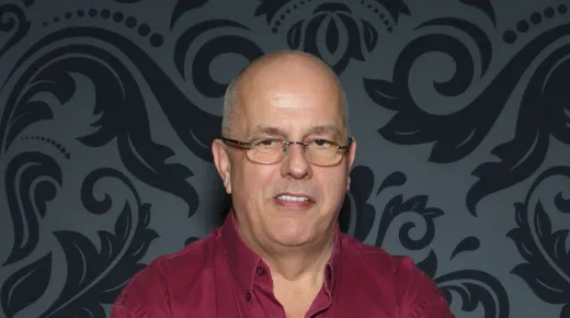 A portrait of an older man wearing glasses and a red collared shirt against a dark patterned wallpaper background.