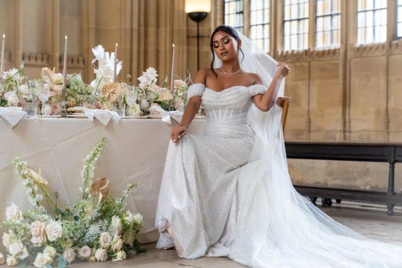 A stunning bride poses gracefully in a beaded off-the-shoulder gown next to an elaborately decorated wedding breakfast table inside a historic hall.