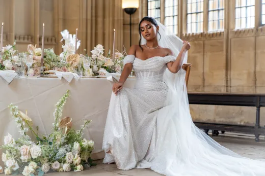 A stunning bride poses gracefully in a beaded off-the-shoulder gown next to an elaborately decorated wedding breakfast table inside a historic hall.