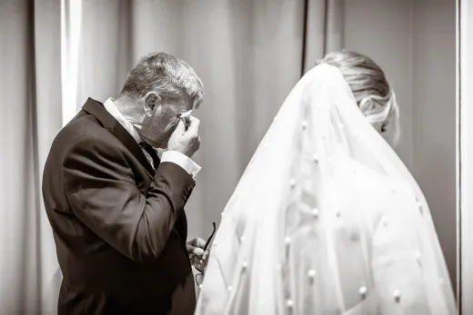 An emotional father of the bride wipes a tear from his eye while sharing a special moment with his daughter, who wears a pearl-embellished veil.