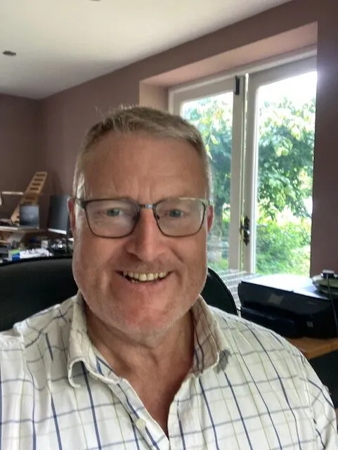 A smiling man wearing glasses and a checked shirt sits indoors near a window.