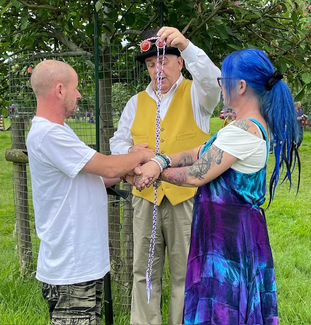A celebrant wearing a top hat with goggles and a yellow waistcoat officiates an outdoor handfasting ceremony in an orchard, wrapping a braided cord around the hands of a couple with vibrant tattoos and blue hair.