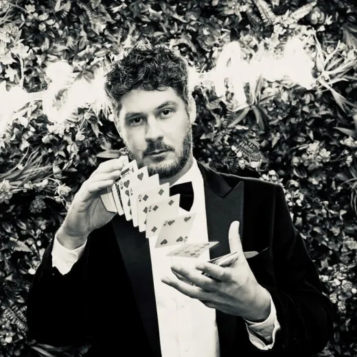 A magician in a black tuxedo performs a card trick in front of a foliage and neon backdrop during a wedding reception.