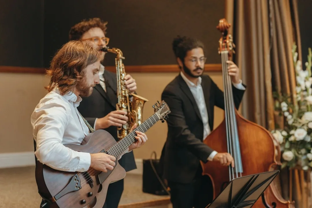 A live wedding jazz band performs indoors, featuring a guitarist in the foreground, a saxophonist, and a double bassist.