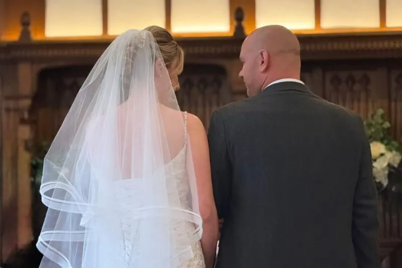 A bride and groom are seen from behind during their civil ceremony inside a wood-panelled room, with the bride wearing a lace gown and long veil and the groom in a grey suit.