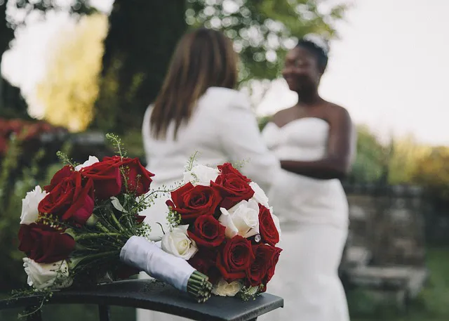 A close-up of a red and white rose bouquet resting on a surface while two brides stand softly blurred in the background.