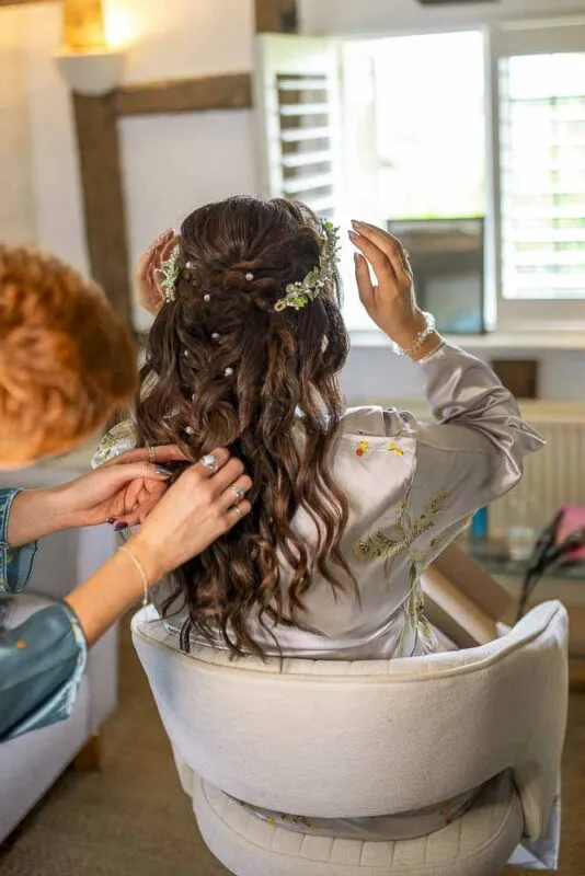 A bride sits in a white armchair facing a bright window while getting her long brown hair styled in soft waves with delicate pearl pins and a floral hair vine during morning preparations.