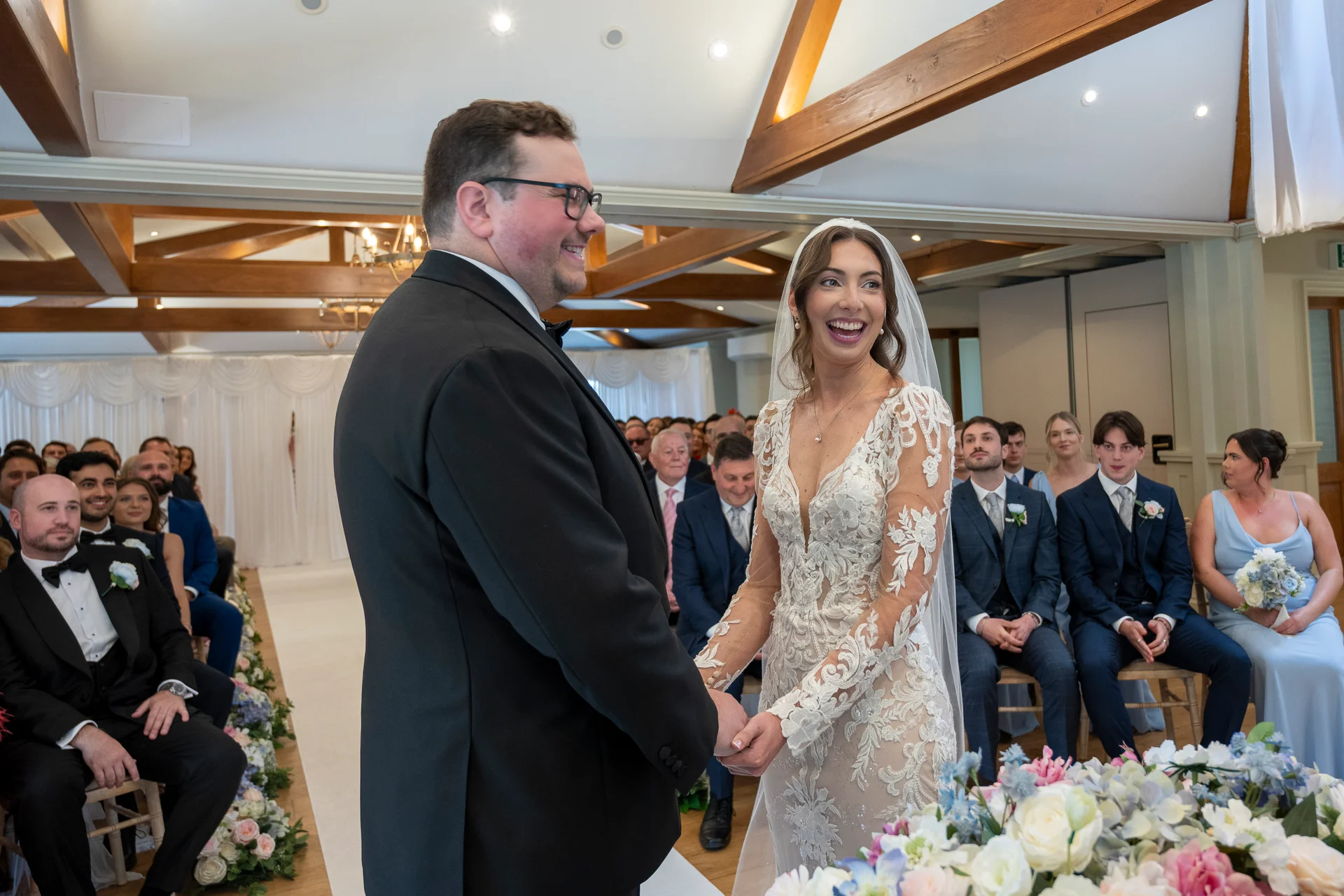 A smiling bride in a lace long-sleeve gown and groom in a black tuxedo hold hands during their light-filled indoor civil ceremony.