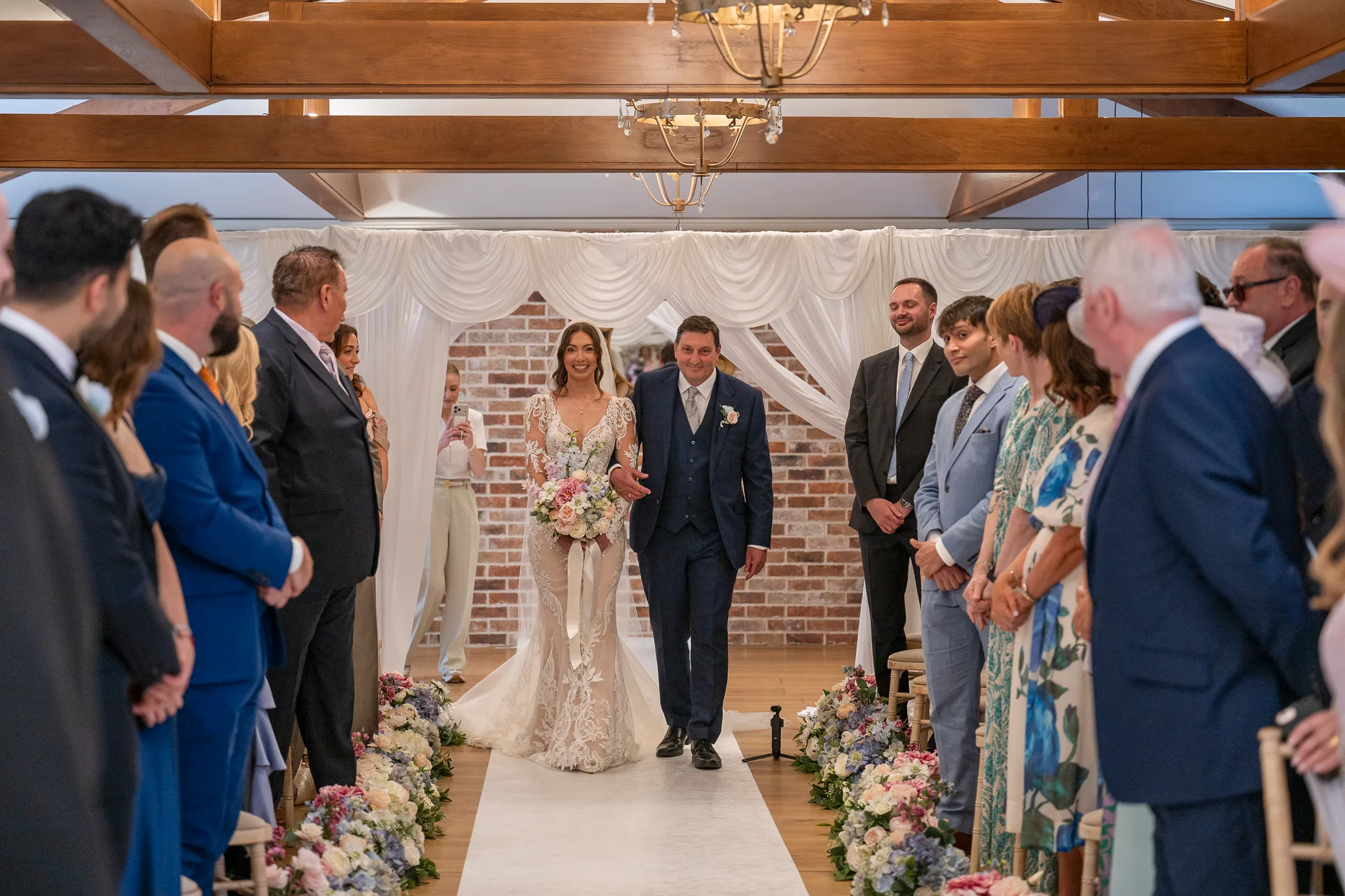 A smiling bride walks down the aisle arm in arm with her father, carrying a delicate floral bouquet inside a venue with exposed wooden beams and floral arrangements lining the walkway.