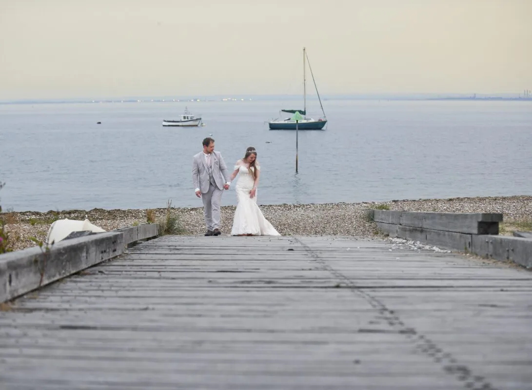 A newlywed couple walks hand in hand along a pebble beach toward a wooden pier with sailboats on the water in the background.