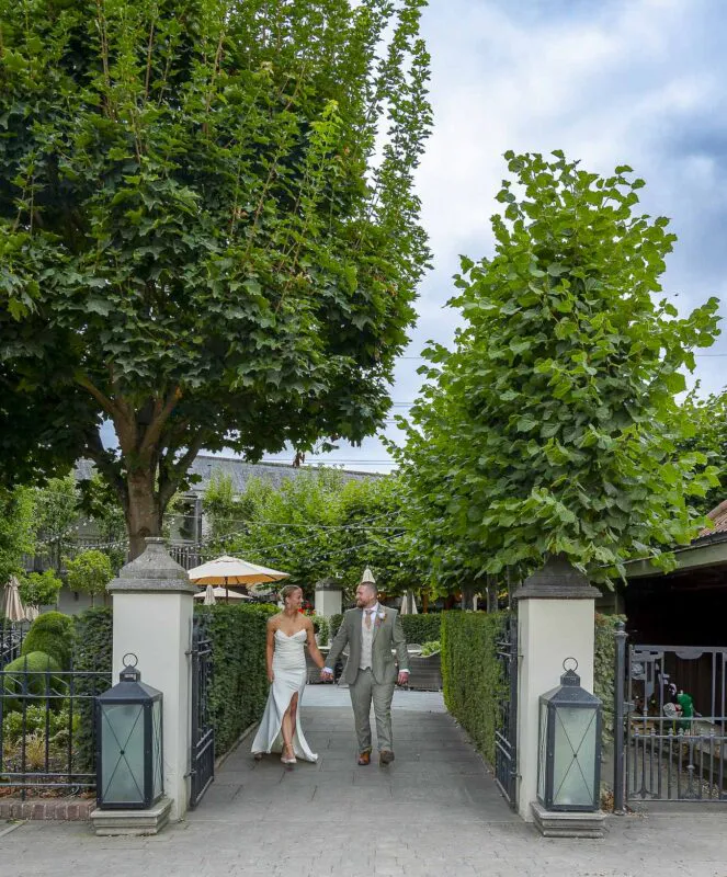 A bride and groom walk hand in hand along a stone pathway framed by stone pillars and lush green trees.