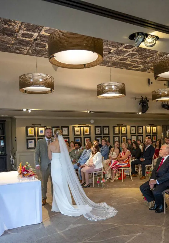 A newlywed couple stands facing each other during their indoor wedding ceremony, with the bride wearing a sleek column dress and long chapel veil while guests watch attentively.
