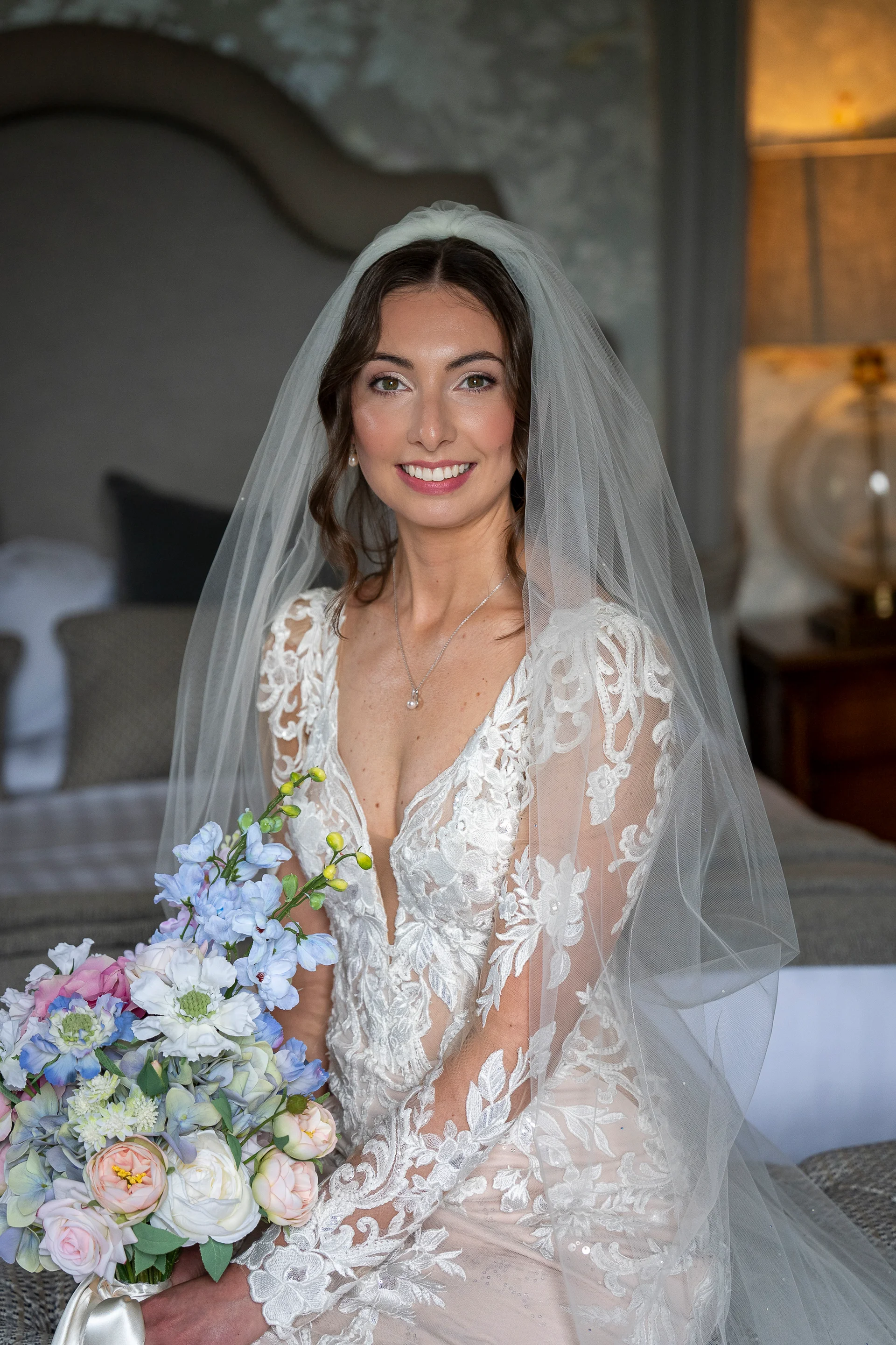 A beautiful bride with a veil and lace gown smiles warmly while holding a colourful pastel bouquet of roses and delphiniums indoors.