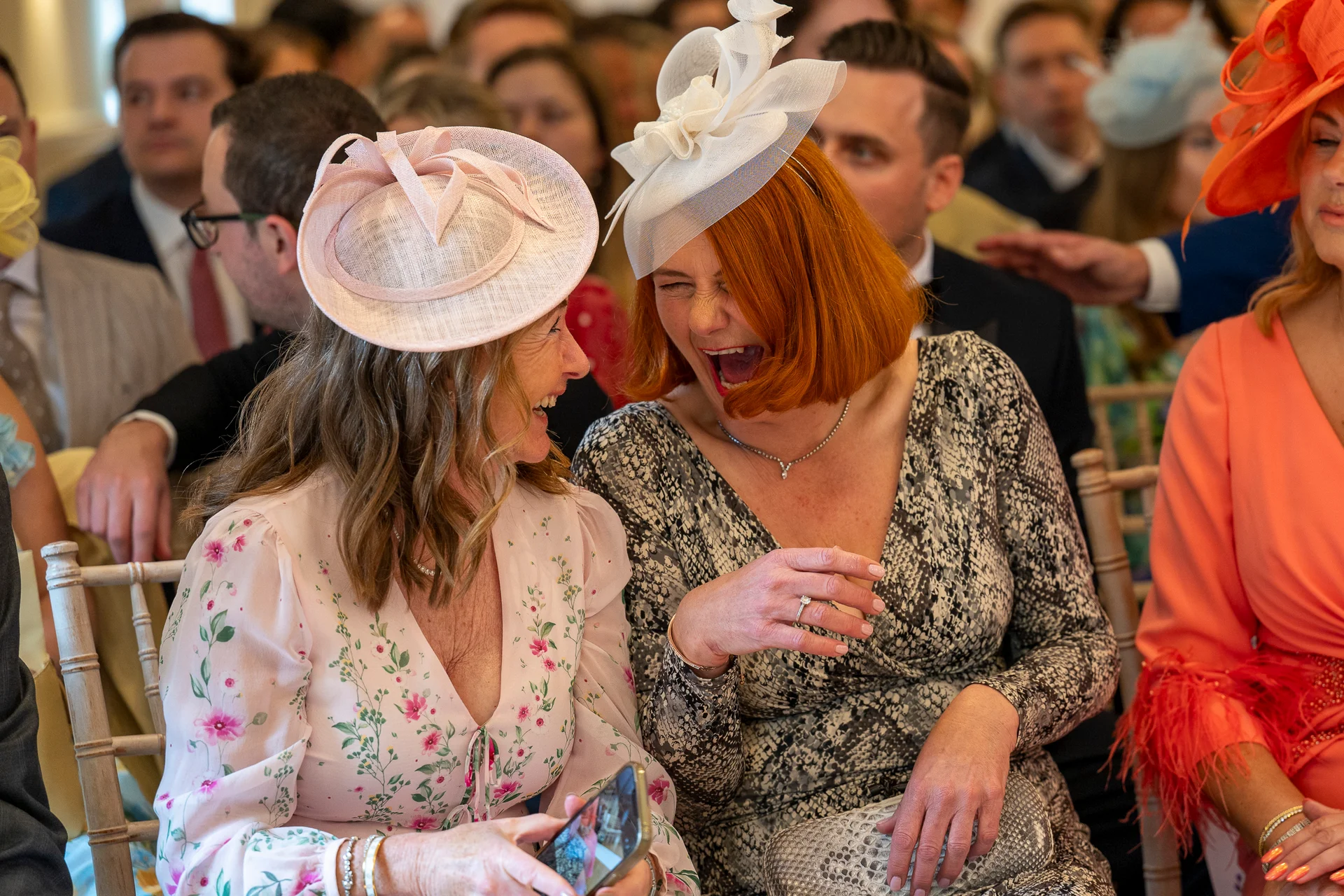 Two wedding guests wearing stylish fascinators share a joyful belly laugh while seated among the crowd during the ceremony.