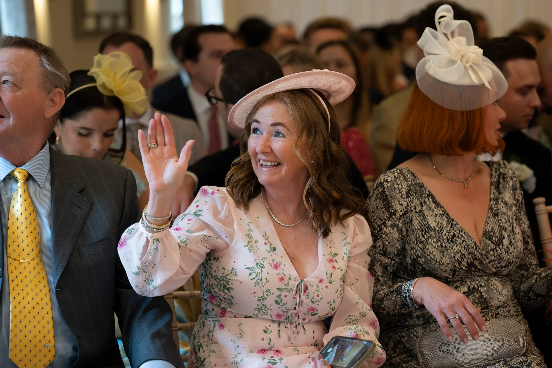 A joyful wedding guest in a floral pink dress and matching hat waves cheerfully while seated among family and friends during the ceremony.