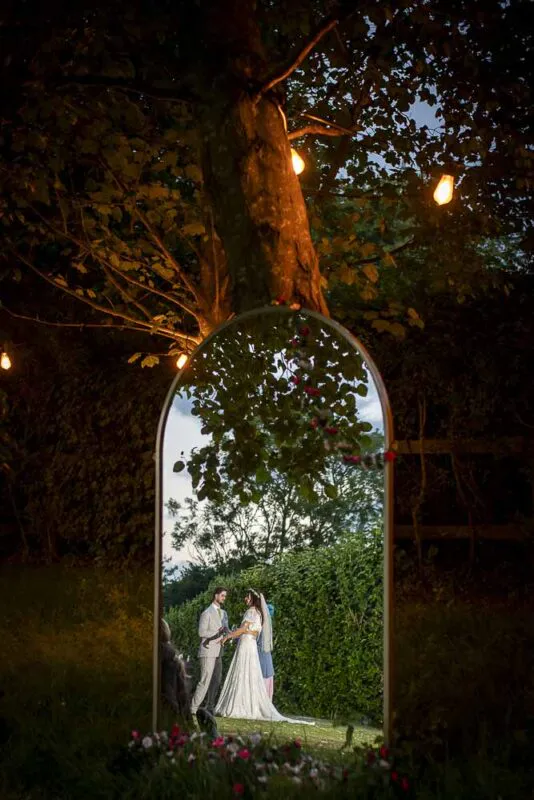 A newlywed couple is captured in a romantic mirror reflection framed by trees and glowing string lights at an evening wedding celebration.