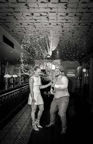 A newlywed couple excitedly spraying champagne inside a dimly lit pub with a distinctive tiled ceiling.