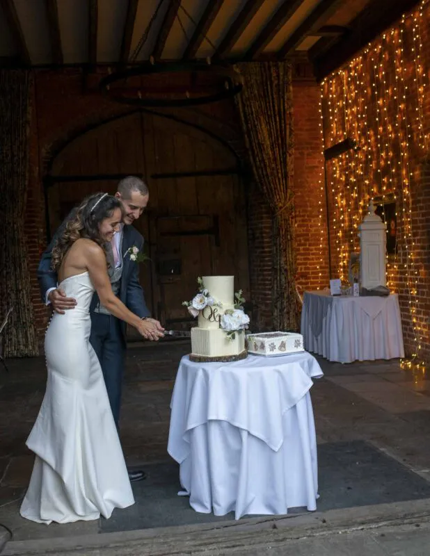 A smiling bride and groom cut their multi-tiered wedding cake inside a rustic barn venue illuminated by a warm fairy light wall.