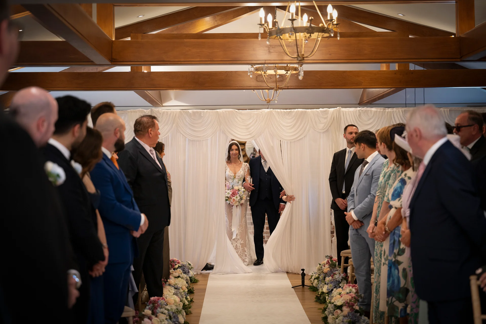 A stunning bride makes her grand entrance down the aisle, holding a delicate bouquet while guests look on inside a rustic barn venue with exposed wooden beams.