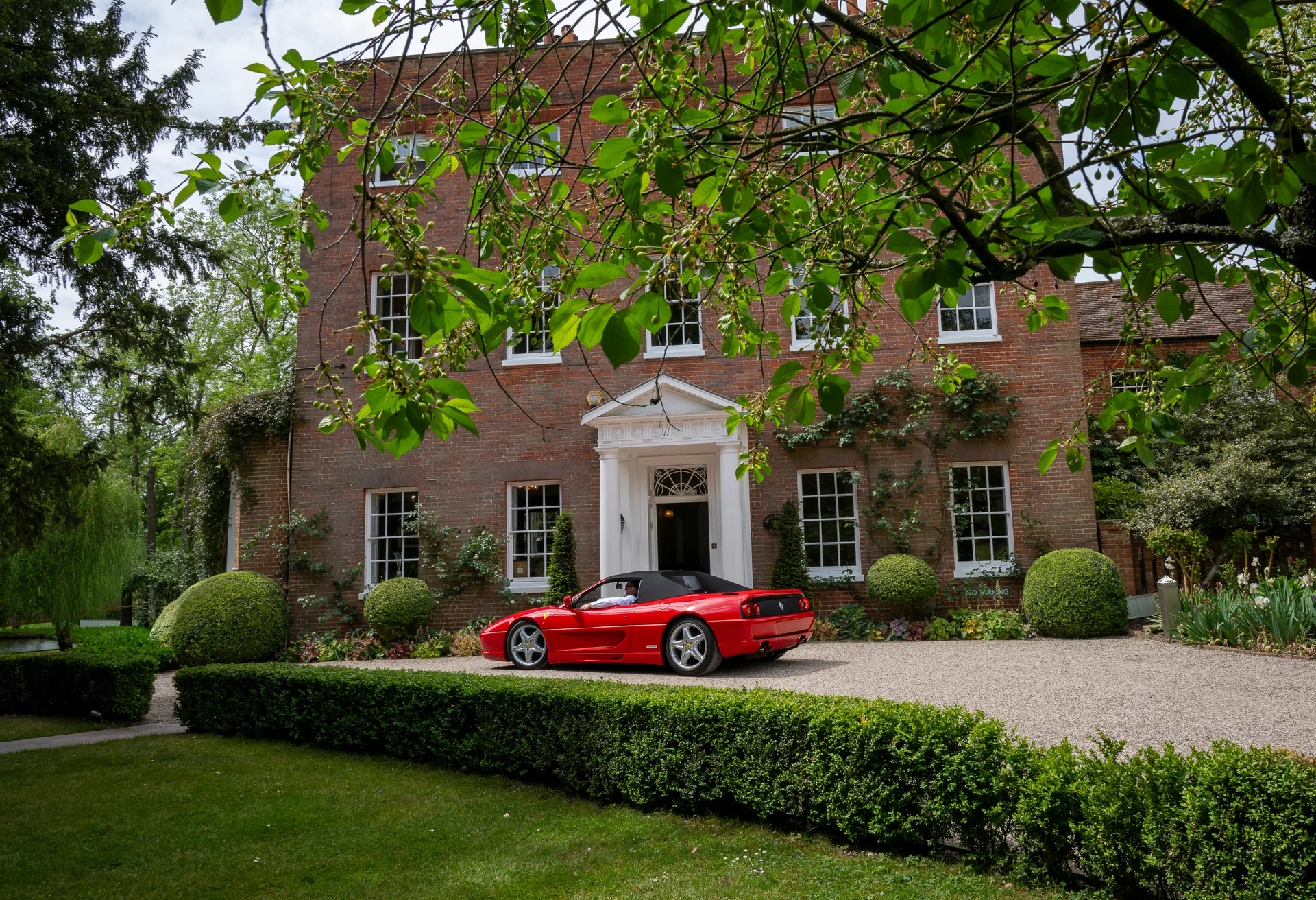 A sleek red sports car parks outside the grand entrance of an imposing red brick country manor house surrounded by manicured topiary and lush gardens.