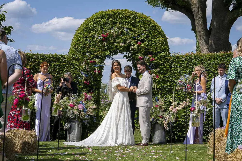 An outdoor wedding ceremony takes place on a sunny day with the bride and groom standing beneath a large green topiary arch while guests watch from hay bale seats.