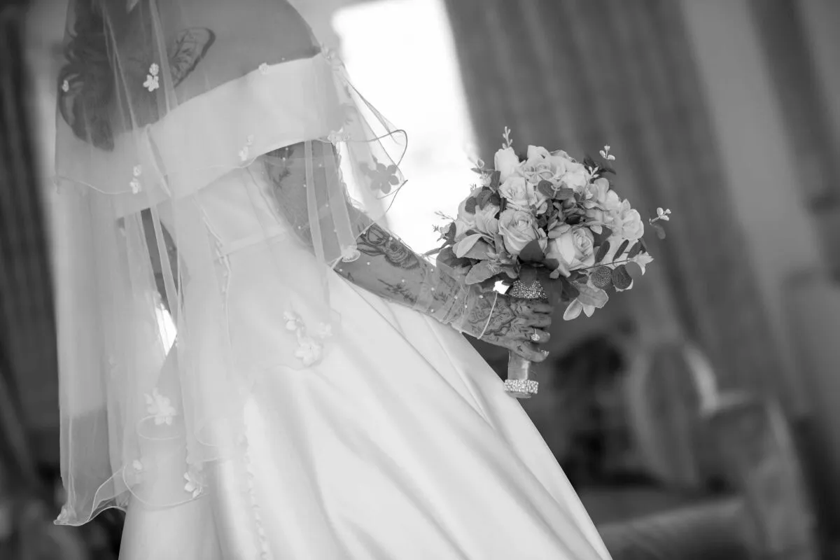 A stunning black and white rear view of a bride wearing an off-the-shoulder satin gown and detailed veil, holding a hand-tied floral bouquet.