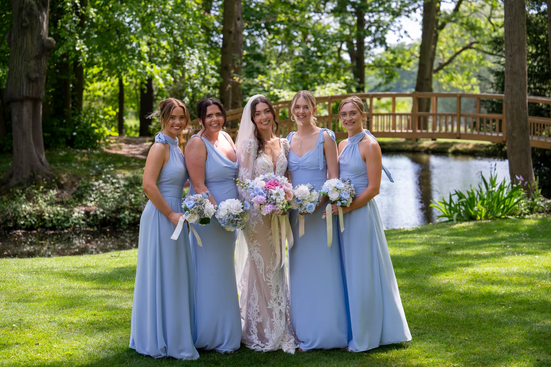 The radiant bride poses outdoors by a riverbank with her four bridesmaids, all wearing matching dusty blue dresses and holding beautiful floral bouquets.