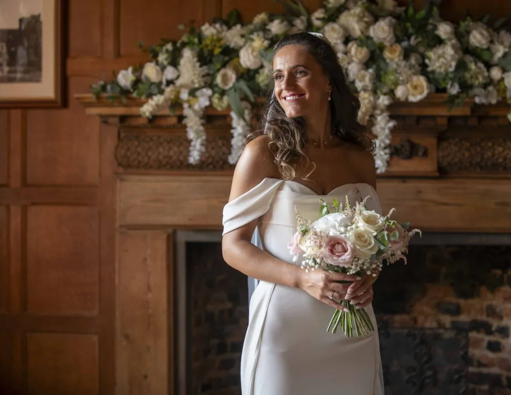 A smiling bride holding a hand-tied bouquet of blush and white roses stands in front of a grand wooden fireplace decorated with a lush floral arrangement.