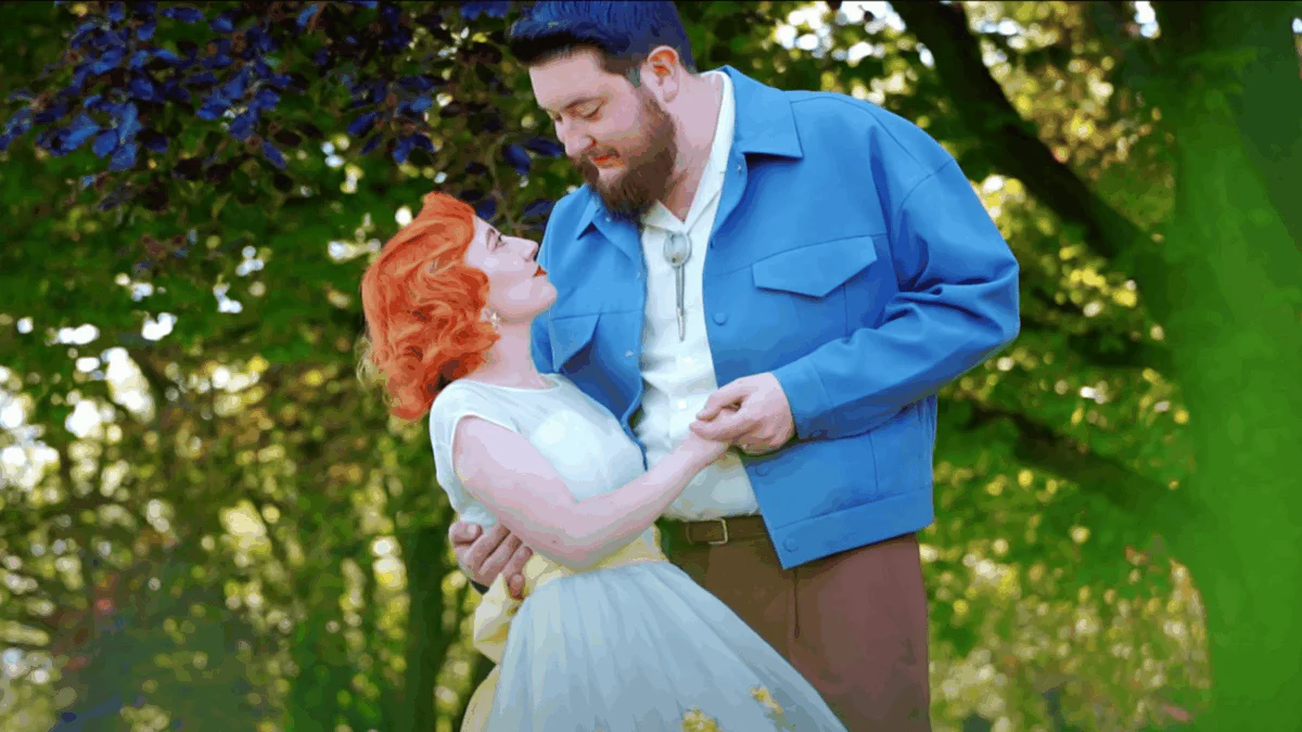 A newlywed couple shares a tender glance in a sunlit woodland setting, with the bride wearing a vintage-style blue dress and the groom in a bright blue jacket.