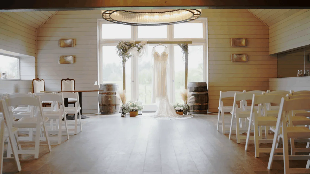 A wedding dress hangs centrally on a wooden ceremony arch in a bright, rustic barn venue featuring rows of white folding chairs.