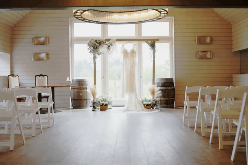 A wedding dress hangs centrally on a wooden ceremony arch in a bright, rustic barn venue featuring rows of white folding chairs.