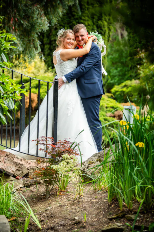 A newlywed couple laugh and embrace on a stone bridge surrounded by lush green garden foliage on their wedding day.