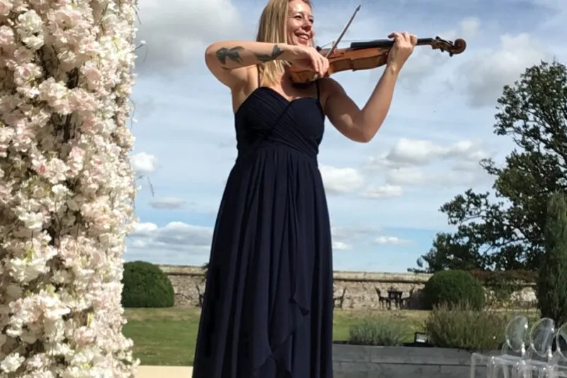 A female violinist wearing a navy blue gown plays her violin outdoors next to a tall floral pillar under a bright blue sky.