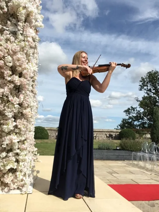 A female violinist wearing a navy blue gown plays her violin outdoors next to a tall floral pillar under a bright blue sky.