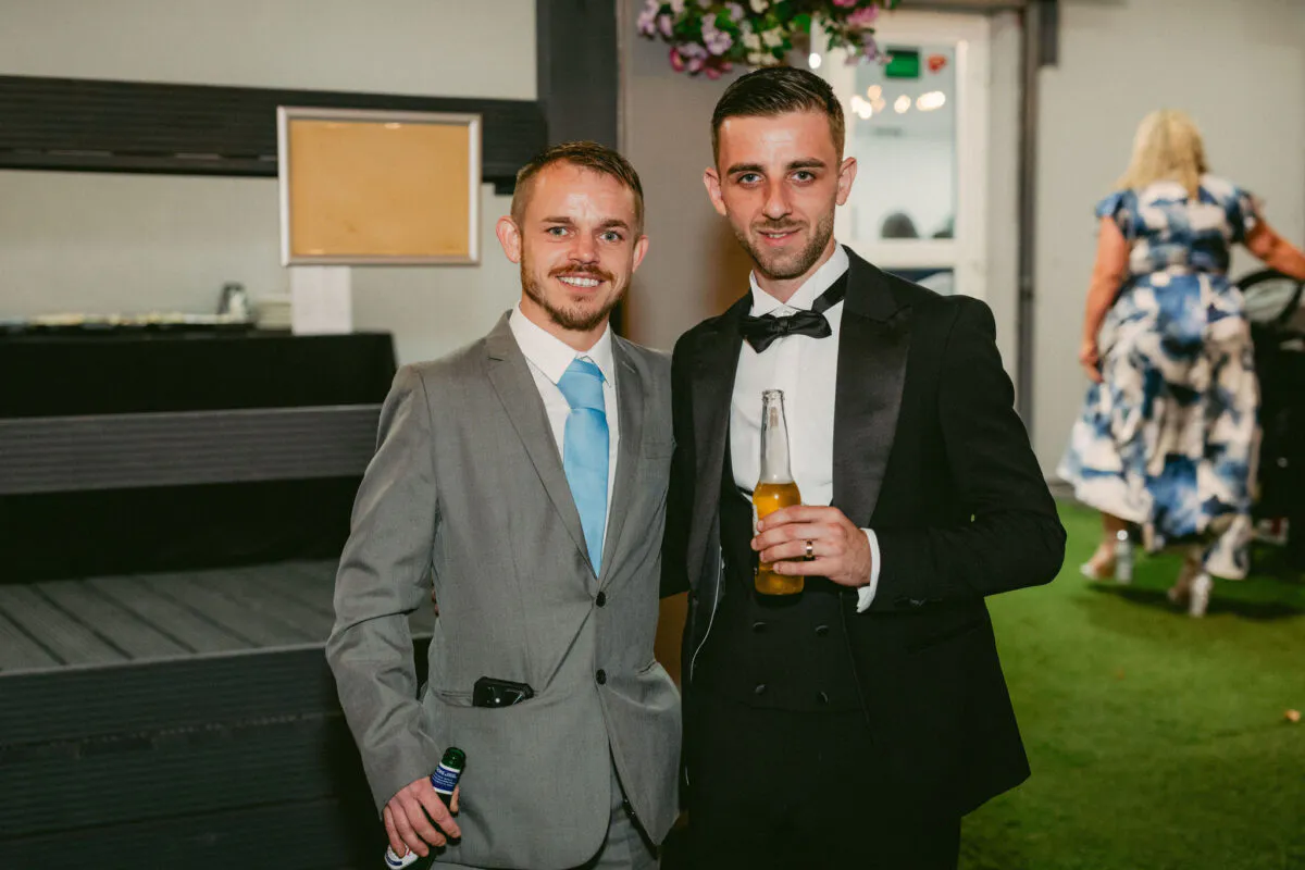 Two grooms in a grey suit and a black tuxedo pose smiling together holding bottled drinks at an evening wedding reception.